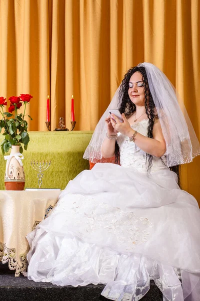 Uma noiva judia em um vestido de casamento branco com um véu senta-se a uma mesa com flores comunicando-se em mensageiros instantâneos aceitando parabéns antes da cerimônia do chuppah. — Fotografia de Stock