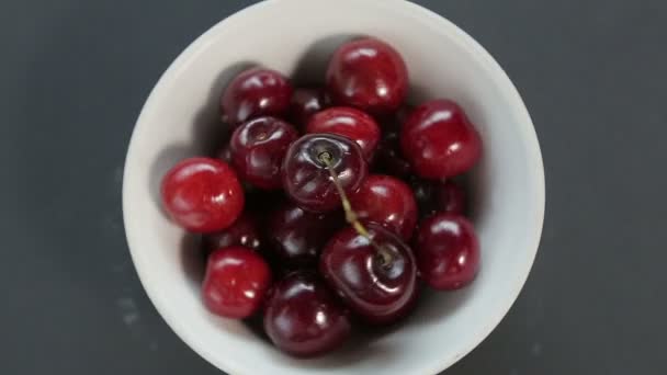 Las cerezas maduras jugosas en un tazón blanco sobre un fondo negro rotan en círculo — Vídeo de stock