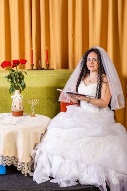 Duvağı olan beyaz gelinlikli Yahudi bir gelin, çiçeklerle dolu bir masada oturur ve törenden önce dua kitabından dualar okur. Dikey fotoğraf