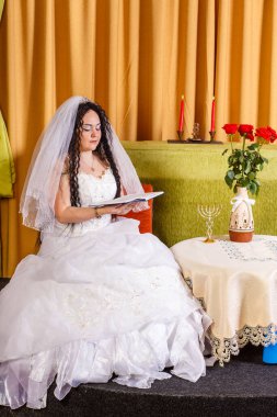 Duvağı olan beyaz gelinlikli Yahudi bir gelin, çiçeklerle dolu bir masada oturur ve törenden önce dua kitabından dualar okur. Dikey fotoğraf