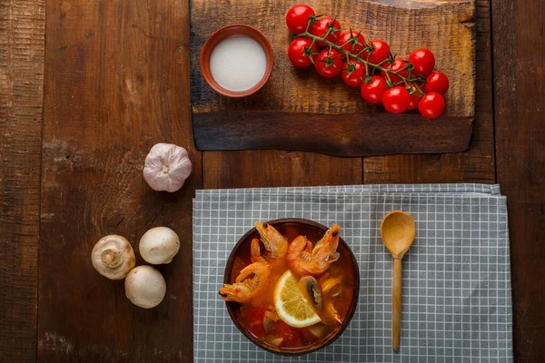 Tom yam soup with shrimps and coconut milk on the table on a checkered napkin next to vegetables and a wooden spoon. — Stock Photo, Image