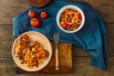 Domatesli deniz kabuğu makarnası, tavuk budu ızgarada pişmiş, mavi peçetede, çatal ve sebze salatasının yanında..