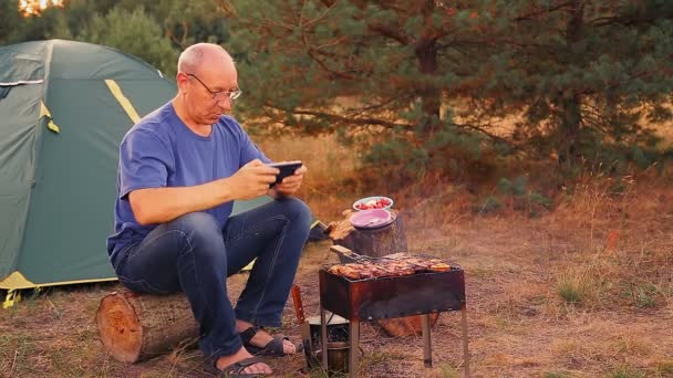 Kamp ateşi çadırının yanında bir adam yemek hazırlıyor ve akıllı telefonuyla oynuyor. — Stok video