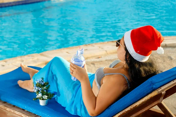 Женщина в шляпе Санты выпивает коктейль в шезлонге с ноутбуком у бассейна во время рождественских каникул возле елки. — стоковое фото
