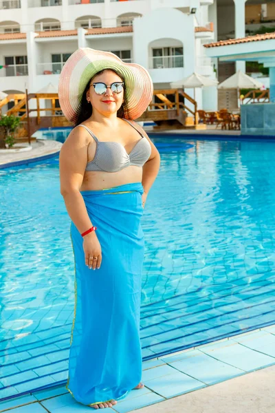 Een lachende brunette in een badpak en een hoed staat bij het zwembad. — Stockfoto