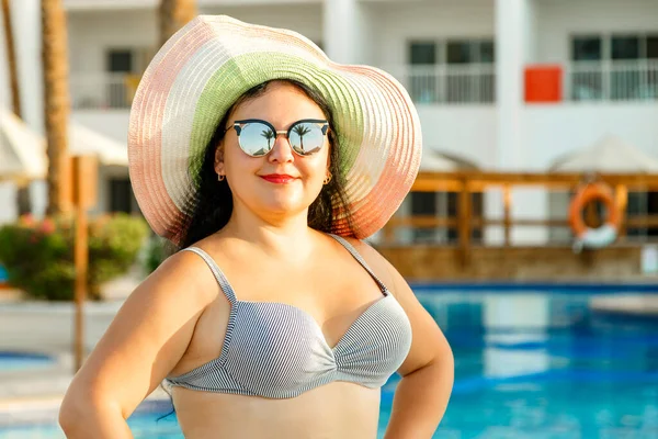 Havuz kenarında pareo mayosu ve şapkası olan gülümseyen bir kadın. Yatay fotoğraf