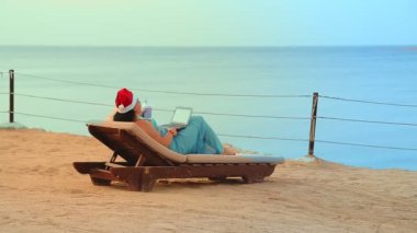 Deniz kıyısında Noel Baba şapkalı esmer bir kadın kokteyl içer ve yeni yılda video bağlantısıyla iletişim kurar.