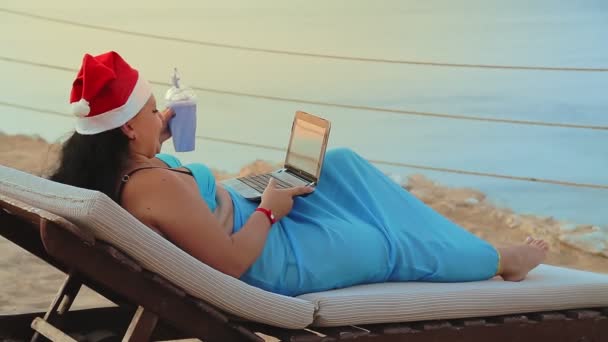 Een vrouw aan zee in een ligstoel in een badpak en een kerstmuts drinkt een cocktail en feliciteert vrienden met het nieuwe jaar vanaf de laptop — Stockvideo