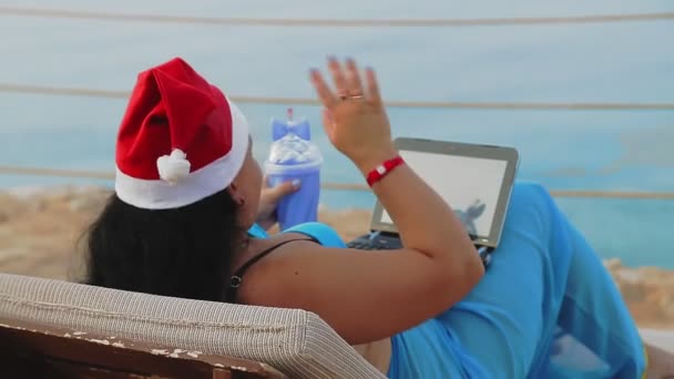 Deniz kıyısında Noel Baba şapkalı esmer bir kadın kokteyl içer ve yeni yılda arkadan video bağlantısıyla haberleşir.. — Stok video