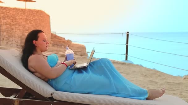 En kvinna i en pareo i en solstol på stranden dricker en cocktail och sms:ar på en bärbar dator — Stockvideo