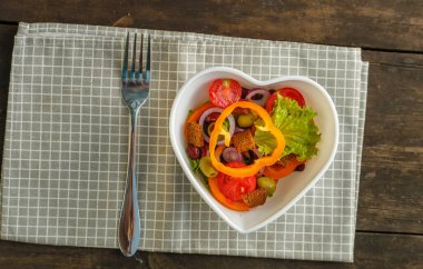 Sebze salatası, kalp şeklinde bir tabakta, bir peçetenin üzerindeki ahşap masada. Yatay fotoğraf