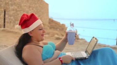 Kumsalda Noel Baba şapkalı esmer bir kadın kokteyl içer ve dizüstü bilgisayarından arkadaşlarını tebrik eder.