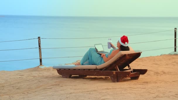 Een brunette vrouw aan de kust in een ligstoel in een kerstman hoed drinkt een cocktail en communiceert via video link in het nieuwe jaar. — Stockvideo