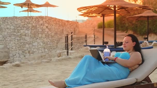 한 여자 가 바닷가에 있는 선더러 너에 누워 칵테일을 마시고 노트북 컴퓨터를 가지고 일하며 측면에서 촬영을 하고 있습니다. — 비디오