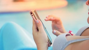 Havuzun yanındaki güneş pansiyonunda mayo giymiş bir kadının elleri akıllı telefondan iletişim kuruyor.