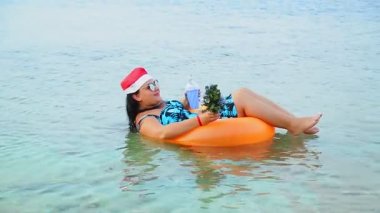 Noel Baba şapkalı ve güneş gözlüklü esmer bir kadın Noel tatilinde elinde bir Noel ağacıyla denizde yüzer ve kokteyl içer..