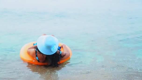 Uma mulher morena de chapéu azul flutua em um círculo no mar e olha para a distância — Vídeo de Stock