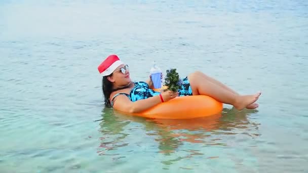 Una donna bruna con un cappello da Babbo Natale e occhiali da sole durante le vacanze di Natale nuota in cerchio nel mare con un albero di Natale in mano e beve un cocktail. — Video Stock