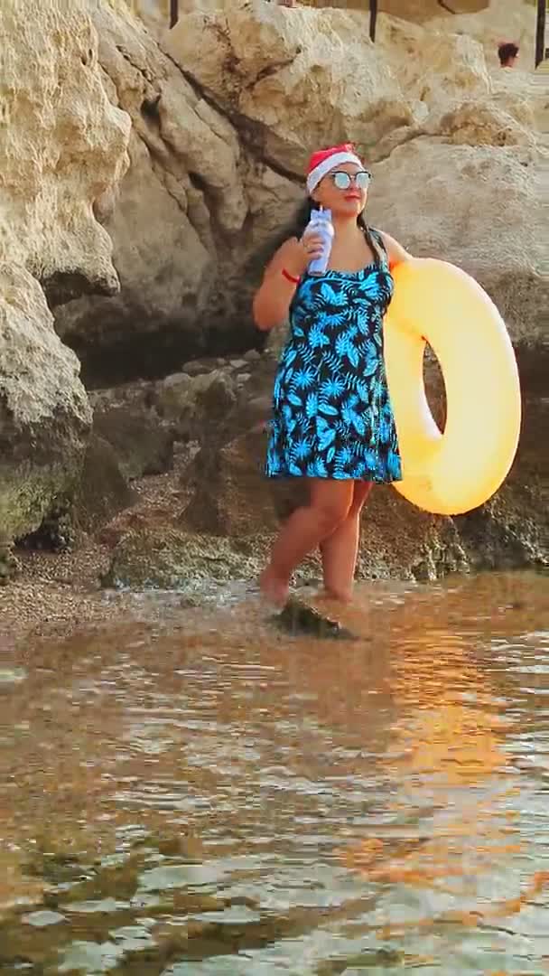 Una mujer morena en un sombrero de santa claus y gafas de sol en las vacaciones de Navidad se encuentra en el borde del mar con un círculo de natación — Vídeos de Stock