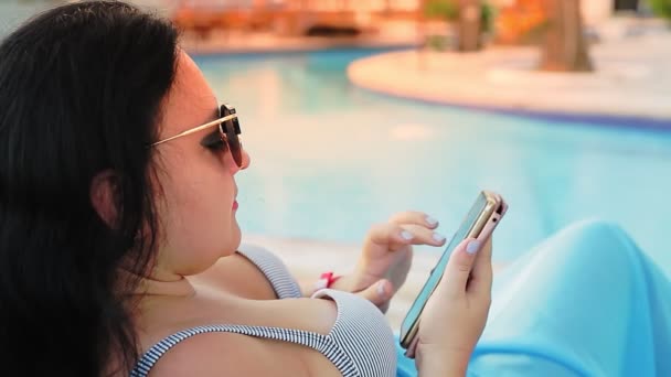 수영장 근처의 선크림 라운지에서 수영복을 입고 선글라스를 끼고 있는 갈색 머리의 여성 이 스마트폰으로 대화를 나눈다. 측면에서 촬영 — 비디오