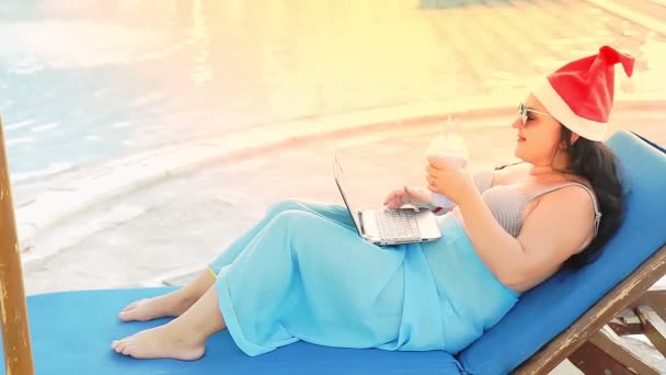 圣诞假期期间，一位戴着圣爪帽躺在游泳池边晒太阳的女人通过视频通话和喝鸡尾酒。来自太阳的光芒 — 图库视频影像
