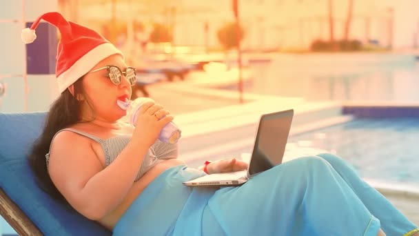 Una mujer en una tumbona junto a la piscina en un sombrero de Santa Claus bebe un cóctel durante las vacaciones de Navidad. deslumbramiento de la luz solar — Vídeos de Stock