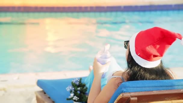 Una mujer en una tumbona junto a la piscina en un sombrero de Santa Claus durante las vacaciones de Navidad bebe un cóctel. deslumbramiento de la luz solar — Vídeos de Stock