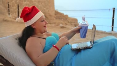 Deniz kıyısında Noel Baba şapkalı bir kadın kokteyl içer ve dizüstü bilgisayardan arkadaşlarını tebrik eder..