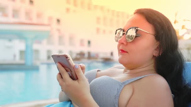 Uma mulher morena em um maiô e pareo e óculos de sol em uma espreguiçadeira perto da piscina se comunica em um smartphone. Luz solar — Vídeo de Stock