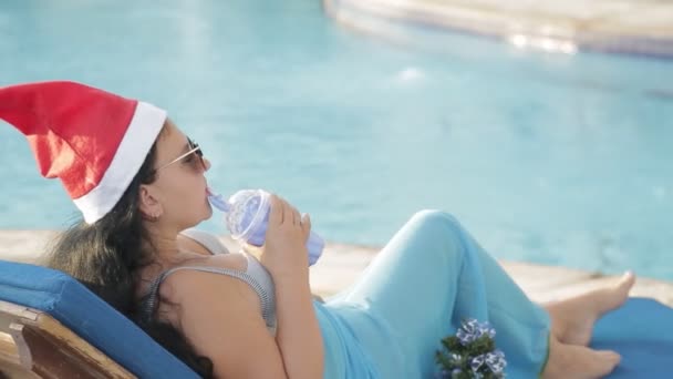 Una mujer en una tumbona junto a la piscina en un sombrero de Santa Claus durante las vacaciones de Navidad bebe un cóctel. deslumbramiento de la luz solar — Vídeos de Stock