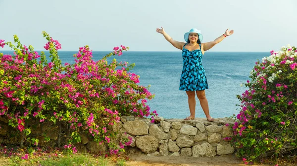 A brunette woman in a blue sundress and hat among blooming bougainvillea stands on a rock against the background of the sea. Horizontal photo