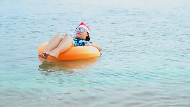 Een brunette vrouw in een kerstman hoed en zonnebril in de kerstvakantie zwemt in een cirkel in de zee en drinkt een cocktail — Stockvideo