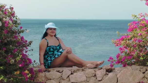 サラファンのユダヤ人女性と帽子は花で飾られた石の壁に座って海を賞賛します — ストック動画