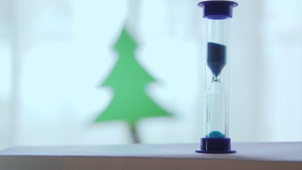 Ampulheta com areia derramando em um fundo branco no fundo o contorno da árvore de Natal — Vídeo de Stock