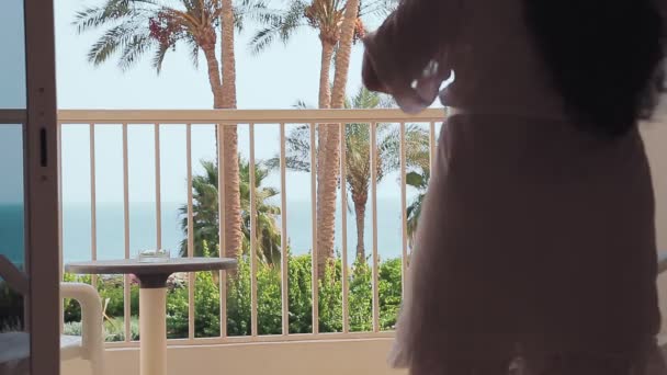 Een vrouw in een wit gewaad opent de gordijnen en stapt uit op een balkon met uitzicht op de zee. Schieten van achteren — Stockvideo