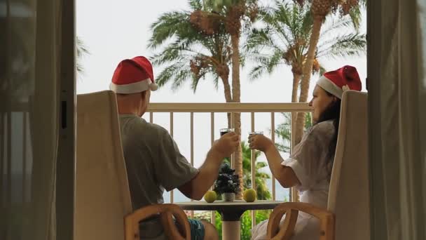 Супружеская пара в шляпах Санта-Клауса празднует Рождество и Новый год на балконе с видом на море на столе маленькая елка — стоковое видео
