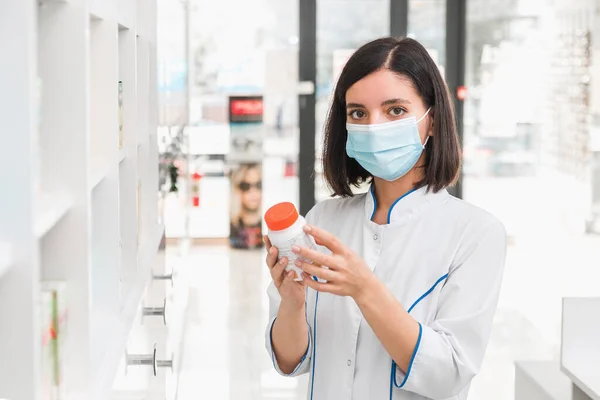 İlaç maskesi takan çekici bir kadın eczacı elinde antiviral ilaçla ilaç kutusu tutuyor.