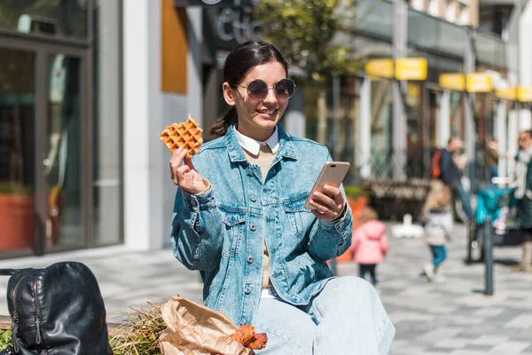 Alegre joven mujer disfrutando de sabrosos gofres en frente de un café en la calle. comida para llevar en la ciudad — Foto de Stock
