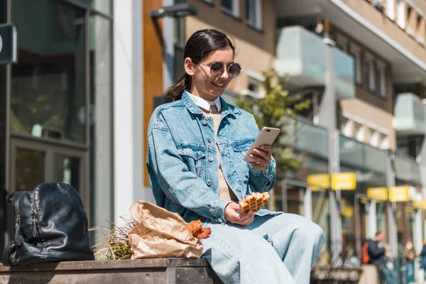 Atractiva chica comiendo en la calle en la ciudad mientras usa el teléfono para calificar la compañía de comida para llevar para delicioso menú — Foto de Stock
