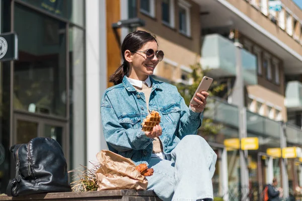 Atractiva chica comiendo en la calle en la ciudad mientras usa el teléfono para calificar la compañía de comida para llevar para delicioso menú — Foto de Stock