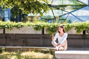 Neşeli etnik köken kız üniversite öğrencisi kampüsün önünde cep telefonuyla mesajlaşıyor.
