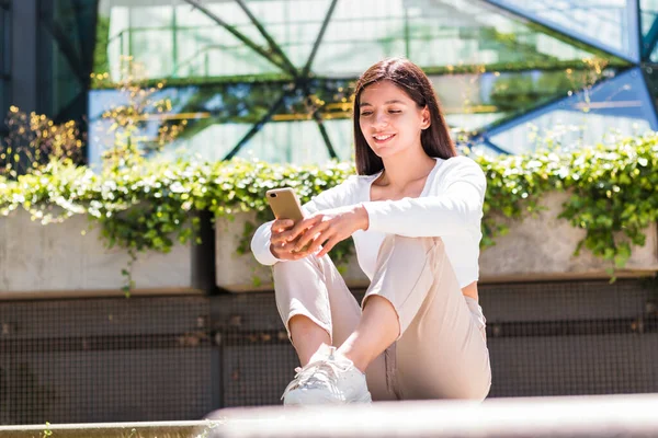 Iloinen etnisyys naispuolinen yliopisto-opiskelija tekstailee matkapuhelimeensa kampuksen edessä — kuvapankkivalokuva