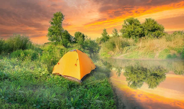 Tienda naranja en la orilla del río en hierba verde bajo el cielo dramático al atardecer — Foto de Stock