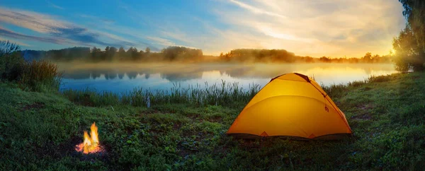 Orangefarbenes Zelt mit Lagerfeuer am Ufer des nebligen Flusses bei Sonnenuntergang — Stockfoto