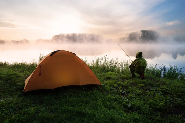 Pescador sentado na costa do lago nebuloso perto de uma tenda laranja — Fotografia de Stock