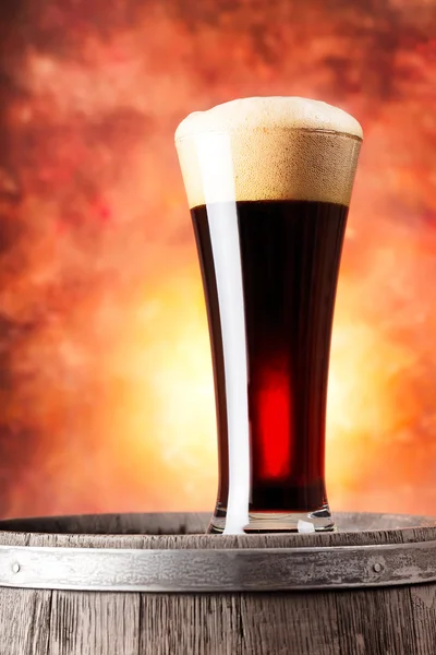 Tall glass of dark beer with foam and wooden barrel