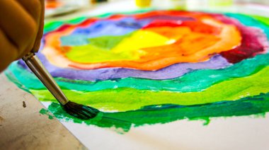 Çocuğun eli beyaz kağıt üzerindeki boyalarla renkli çember çizer.