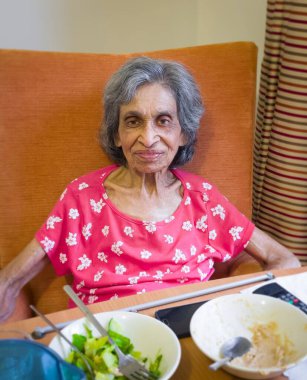 Elderly Indian woman in a care home or nursing home, UK, sitting in a chair eating a meal. Depicts frailty and malnutrition in old age clipart