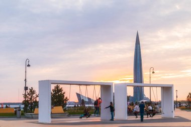 İnsanlar Novokrestovskaya embankmentindeki parkta salıncakta sallanıyor. Hafta sonu Lakhta şehir merkezi günbatımının arka planı. turuncu renk. Rusya, Saint-Petersburg, 14Mayıs 2021