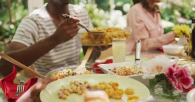 Afro-Amerikalı bir adam bahçede ailesiyle yemek masasında oturuyor, tabağa yemek koyuyor, ağır çekimde..